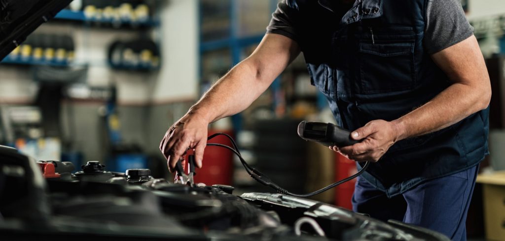 Mid adult mechanic using diagnostic tool while checking voltage of a car battery in auto repair shop.